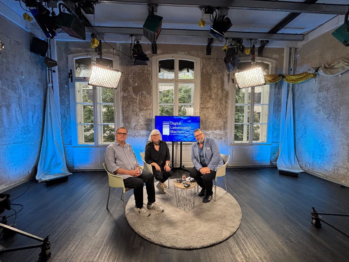 Talkshow „Digital. Liebenswert. Machen.“ im Studio von h1 – drei Personen sitzen auf Stühlen vor einem Bildschirm mit dem Sendungslogo