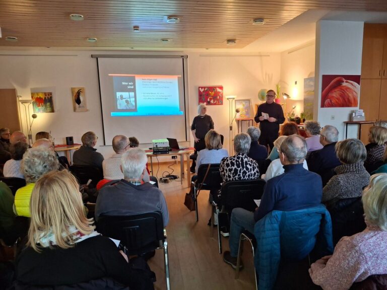 Cornelia Bauer und Hans Vogl stehen vor einer Leinwand mit der Folie „Wer wir sind“, davor ein voll besetzter Raum