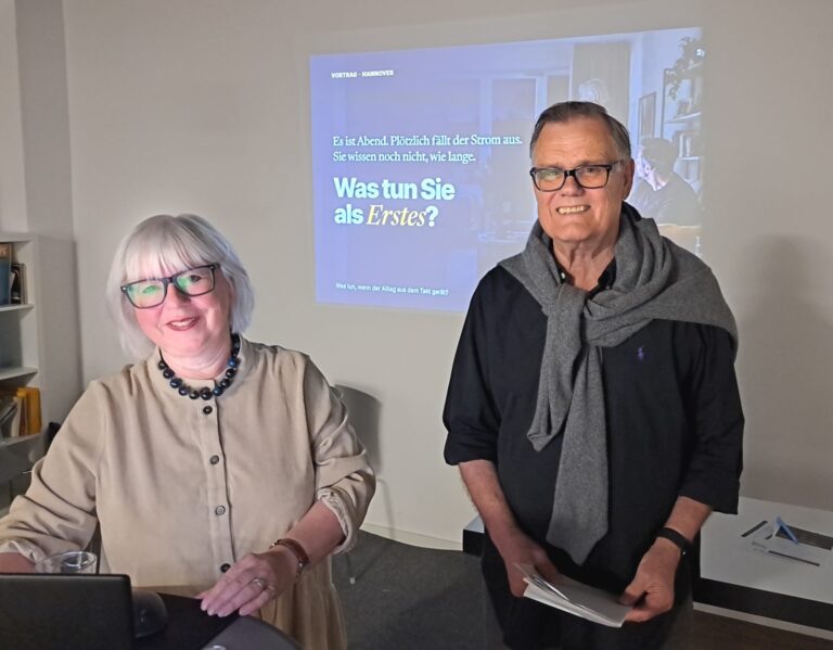 Cornelia Bauer und Hans Vogl stehen lächelnd vor einer Leinwand mit der Frage „Was tun Sie als Erstes?“ zum Thema Stromausfall