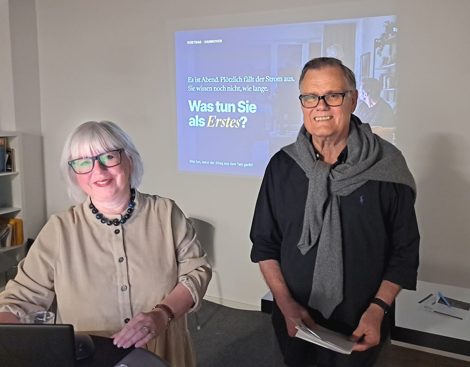 Cornelia Bauer und Hans Vogl stehen lächelnd vor einer Leinwand mit der Frage „Was tun Sie als Erstes?“ zum Thema Stromausfall